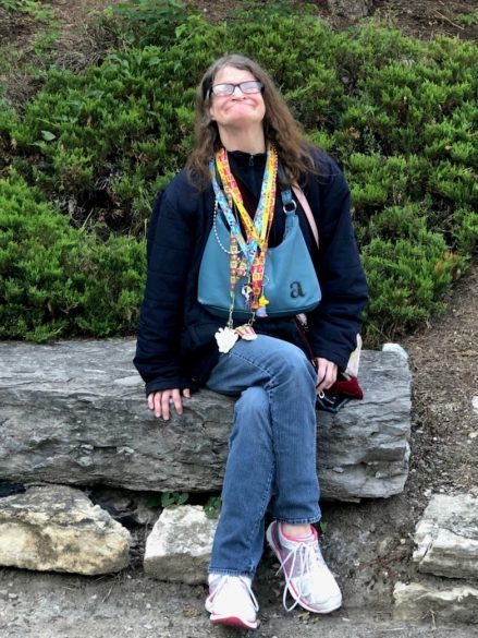 Angee, a woman with long brown hair and eyeglasses wears a navy blue sweater, denim blue jeans, and pink tennis shoes smiles and poses while sitting on a large rock.