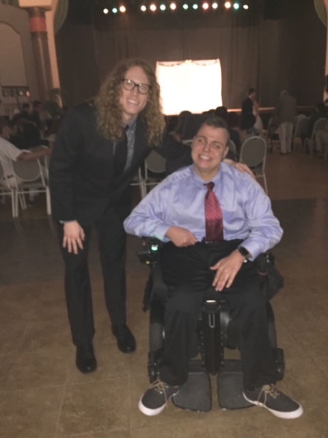 Two men, both dressed in shirt and ties smile for the camera. One of the men has long hair and is standing next to the other that sits in a wheelchair.